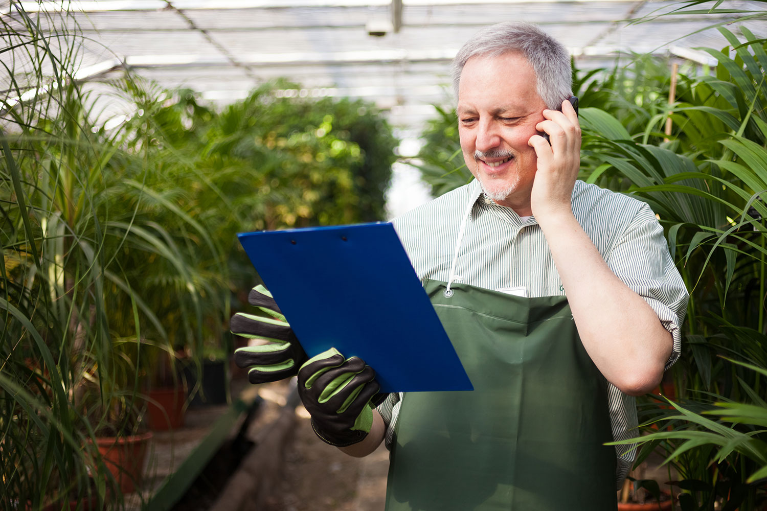 Ein Mitarbeiter des Garten-Centers. Er trägt Gartenhandschuhe und telefoniert am Handy. Er schaut auf ein blaues Klemmbrett vor ihm und lächelt freundlich.